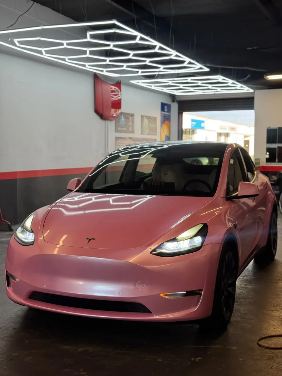 Finished Tesla Model Y wrapped in glossy pink at the Enhanced Automotive shop under hexagon work lights