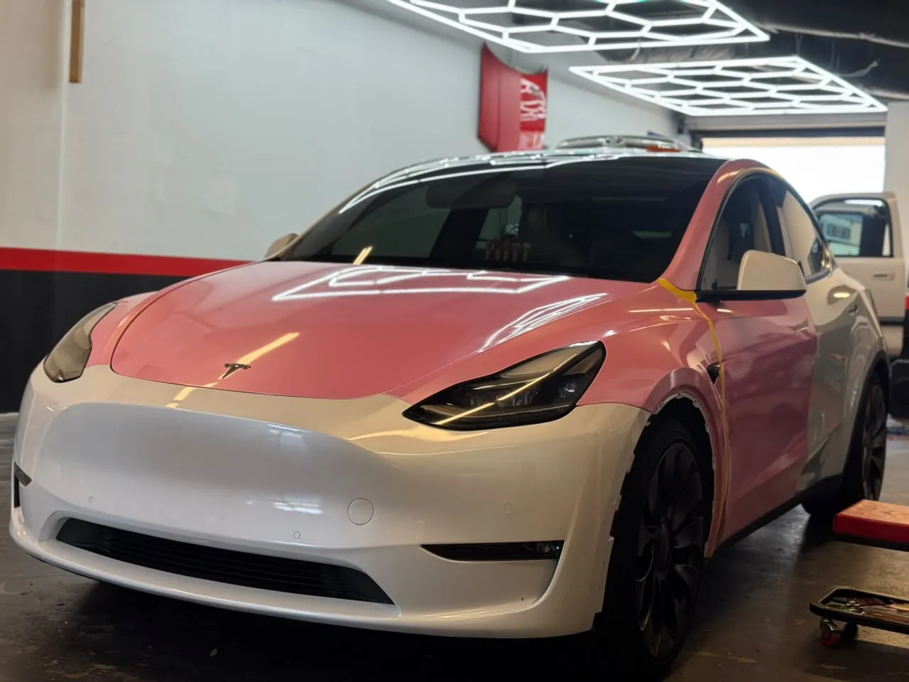 White Tesla Model Y at the Enhanced Automotive shop with pink wrap film applied to the hood and front fender, and yellow tape marking the edge